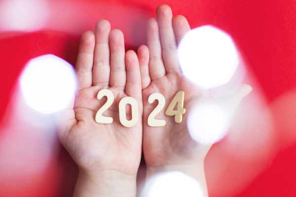 Top view of child hands holding wooden number 2024 on palms on red garland lights bokeh background with copy space. Christmas greeting card, New Year, winter holidays sale and festive mood concept.